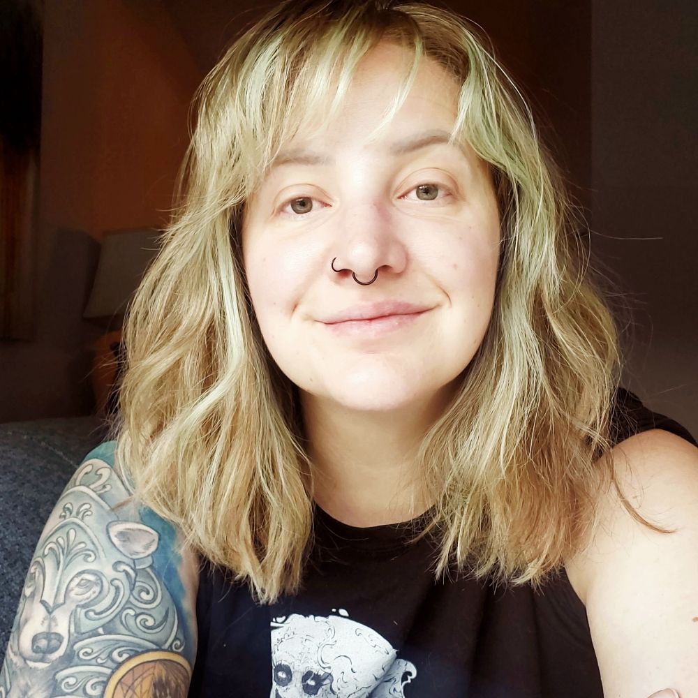 picture of Ash Vale, a white nonbinary person. they have shoulder-length blonde hair and a large tattoo on one arm.